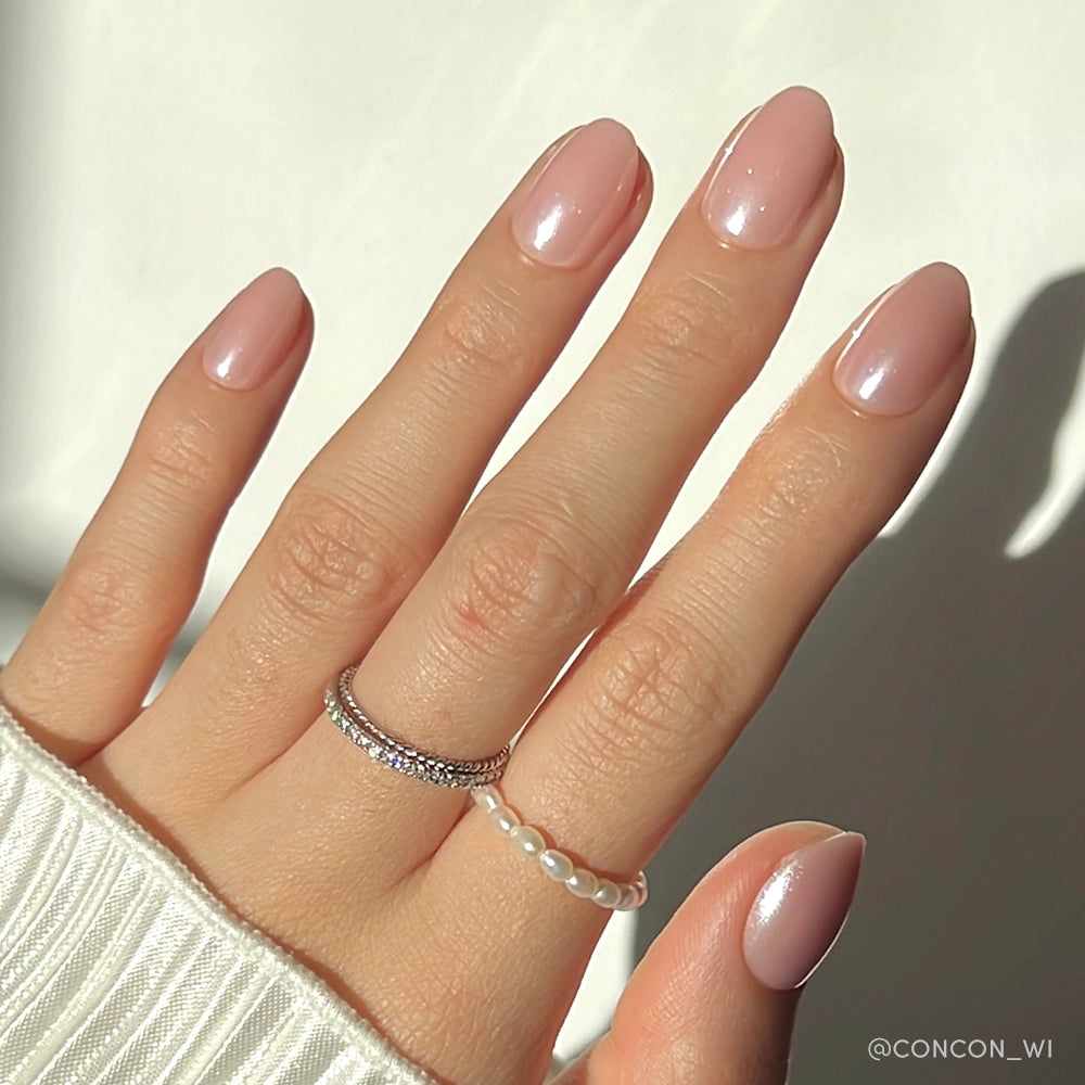 A hand with neatly manicured, short oval nails in glamnetics Pinch Me light pink shade with a glossy glaze, adorned with a silver diamond ring and pearl ring, wearing a white ribbed sweater.